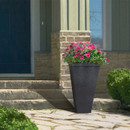 Tall black planter with vibrant pink flowers sits on a stone porch step. Nearby, green bushes and blue door convey a welcoming, peaceful entrance.