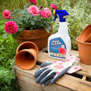 Potted pink roses are beside a bottle of "Rose Clear" spray. Nearby, floral gardening gloves and clay pots are on a wooden surface, evoking a garden setting.