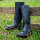 Two navy rubber boots stand against a wooden fence in a grassy field, slightly muddy, with small purple flowers nearby, conveying a rustic, outdoorsy feel.