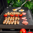 Spatula in use on a hot grill cooking sausages, tomatoes, mushrooms, bacon and fried eggs.