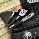 Three-piece barbecue tool set (tongs, spatula and grill brush) resting on a barbecue side shelf.