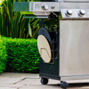 Barbecue with a side-mounted rack storing a pizza stone and griddle plate, outdoors in a garden setting.