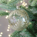 A clear glass bauble with a gold glitter foliage pattern hanging from a Christmas tree with gold string lights in the background.