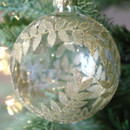 A close up view of a clear glass bauble with a gold glitter foliage pattern hanging from a Christmas tree.
