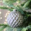 A grey geometric bauble with a diamond pattern, hanging from a Christmas tree with gold string lights in the background.