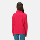 A person with long brown hair, wearing a bright pink fleece jacket and blue pants, is shown from the back against a plain gray background.