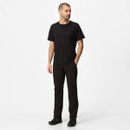 A man stands against a white background, wearing a black t-shirt, black pants, and black shoes, with a neutral expression and his left hand in a pocket.