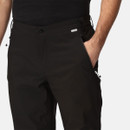 A person wearing black trousers and a black shirt stands against a plain background. One hand is casually placed in a pocket, suggesting a relaxed posture.