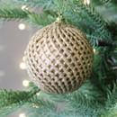 A gold glitter bauble with a geometric diamond pattern, hanging from a Christmas tree with gold string lights in the background.