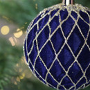 A close-up of a deep blue Christmas ornament with a silver lattice pattern, hanging on a green tree. Soft, warm lights are blurred in the background.