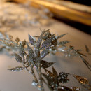 Close-up of glitter-covered leaves on a branch, placed on a smooth surface. The background is softly blurred, creating a festive and elegant atmosphere.