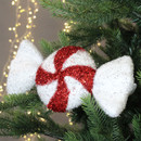 A glittery red and white peppermint candy ornament, shaped like wrapped candy, hangs on a green Christmas tree branch with soft, twinkling lights.