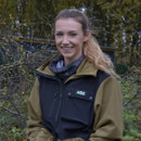 Person in a beige jacket and black vest standing outdoors with trees in the background.