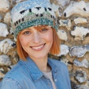 Smiling person with short hair wearing a knitted beanie featuring sheep patterns. Dressed in a denim shirt, they stand against a stone wall.