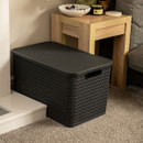 Charcoal woven plastic storage basket with lid placed on a carpeted floor beside a wooden side table, modern home décor storage solution for living room organisation.