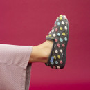 A person wearing a grey Lunar slipper with a fluffy texture and colourful polka dots in pastel hues (blue, pink, white, yellow, and green), raising their foot against a bold magenta background. The person is dressed in light mauve ribbed trousers.
