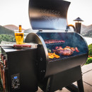 A smoker grill cooking various meats with a beverage on top, set against a scenic backdrop.