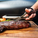 A hand using kitchen shears to cut a piece of perfectly cooked, smoked meat on a cutting board.