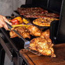 BBQ turner lifting a whole roasted chicken from a smoker grill onto a wooden cutting board, with grilled meats and vegetables in the background.