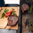A hand holds a grilling tool, flipping juicy steaks and colorful vegetables on a barbecue grill under bright sunlight.