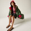 Young girl in a camo jacket with pink lining and a matching pink hat sits on a white block, wearing a pink shirt, black shorts, and flip-flops. Relaxed vibe.