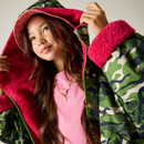 Young person wearing a green camo jacket with pink furry lining, adjusting the hood. They have long hair, a pink shirt, and a necklace, looking content.