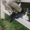 Person wearing black sneakers and beige pants sits on a white wooden fence. Lush green grass and water reflection are visible in the background, creating a serene atmosphere.
