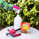 A person wearing gloves pours weed killer into a white spray bottle. Nearby, a red and black nozzle and a Weedol box are on the table. Green leaves form the background.