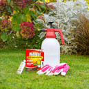 Gardening tools on green grass with vibrant plants in the background. Items include a Weedol box, spray bottle, pink gloves, and gardening tube.