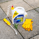 White container of Weedol weed killer, with a yellow battery-powered sprayer, set on stone patio. Yellow garden gloves lie beside a small green weed.