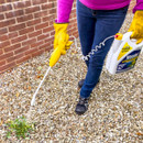 Person in a pink top, jeans, and yellow gloves uses a weed killer sprayer on gravel by a brick wall. The mood is practical and focused.
