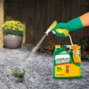 A gloved hand sprays a weed in a gravel area with a green and yellow Roundup weed killer bottle. A potted plant is visible in the background.