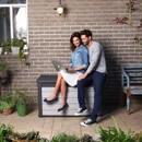 Couple sitting on a grey garden storage box with a laptop in a patio garden setting.