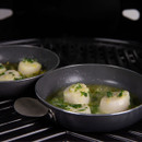 Two pans on a grill, each containing round dumplings in a green herb-infused broth, garnished with fresh herbs.