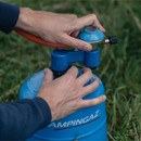 A person's hands attach a regulator to a blue Campingaz gas canister in a grassy outdoor setting.
