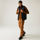 Man smiling in casual outdoor attire; wearing a black cap, black quilted vest, brown long-sleeved shirt, brown cargo pants, and black boots. Neutral background.