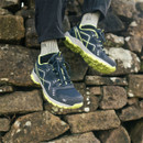 Man wearing navy and lime waterproof hiking trainers with breathable mesh panels, sitting on a stone wall outdoors.