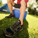 Man sitting outdoors in the sun wearing grey and red walking trainers paired with shorts and black socks.