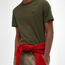 A person wears an olive green t-shirt with a small logo and tan pants. A red jumper is tied around the waist, conveying a casual, stylish look.