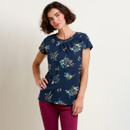 Woman in a floral top and maroon pants standing against a plain background.