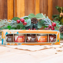 Gift box with five jars of chutney and pâté on a wooden table. Holiday greenery with red berries and pinecones in the background; warm, festive vibe.