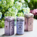 Three ornate tins of English tea, labelled Afternoon, Earl Grey, and Breakfast, are arranged on a table. Vibrant hydrangeas in the blurred background.