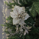 Elegant white and gold flower ornament with glittery leaves decorates a green Christmas tree branch. Soft, warm fairy lights glow in the background.