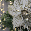 Close-up of a white poinsettia ornament with gold glitter accents on a Christmas tree, surrounded by warm, blurred fairy lights, creating a festive and elegant mood.