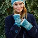 A smiling woman in a blue coat wears matching blue and white knitted hat and fingerless gloves, against a leafy background. A cosy, cheerful vibe.