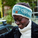 A person smiling warmly, wearing a blue patterned headband and a black coat over a white turtleneck, with a blurred, green outdoor background.