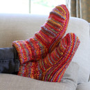 Cosy, colourful knitted slippers with a striped pattern in red, orange, and purple are worn by a person resting on a beige couch, conveying warmth and relaxation.