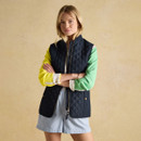 A person stands with arms crossed, wearing a navy quilted gilet over a colourful long-sleeve shirt and light shorts against a beige background.