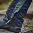 Close-up of a person wearing dark blue overtrousers with a green stripe and rugged black boots standing on wooden planks. The scene is outdoors, suggesting a gardening or farming setting.