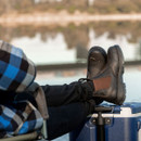 Person in a blue checkered shirt relaxing by a lake, feet up on a cooler, wearing black pull-on work boots with brown elastic sides.